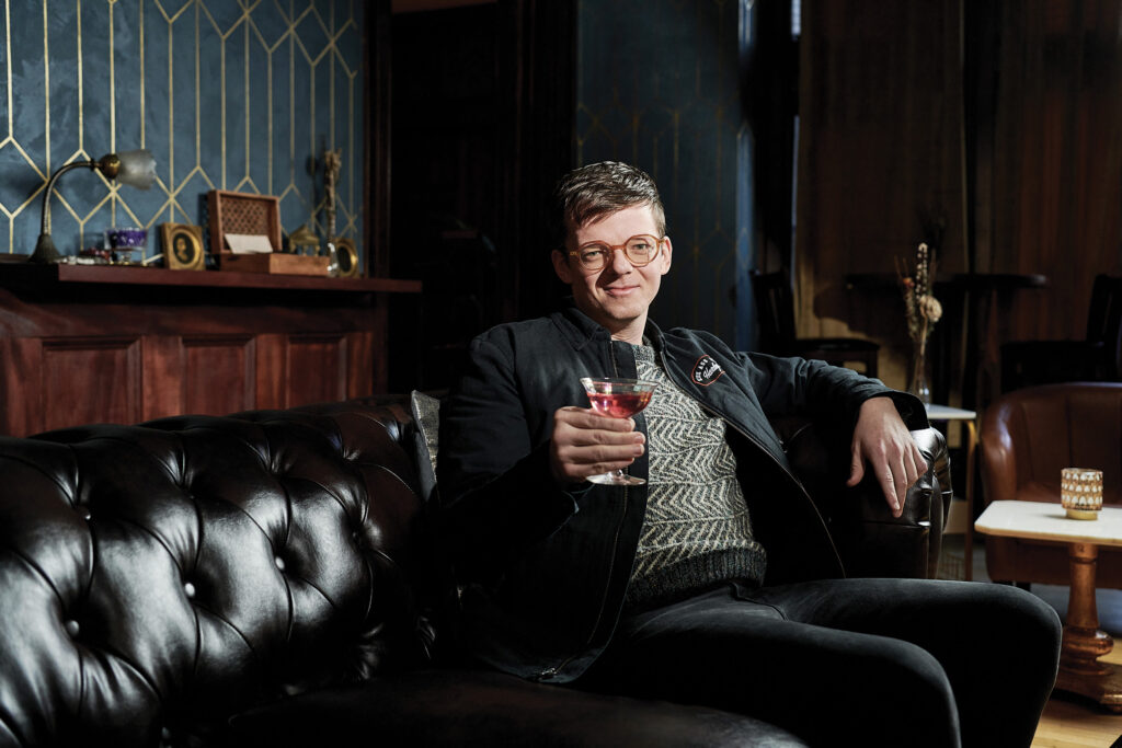 A person wearing glasses and dark clothing sits on a tufted black leather couch in a sophisticated room with wood paneling and decorative objects, holding a drink.