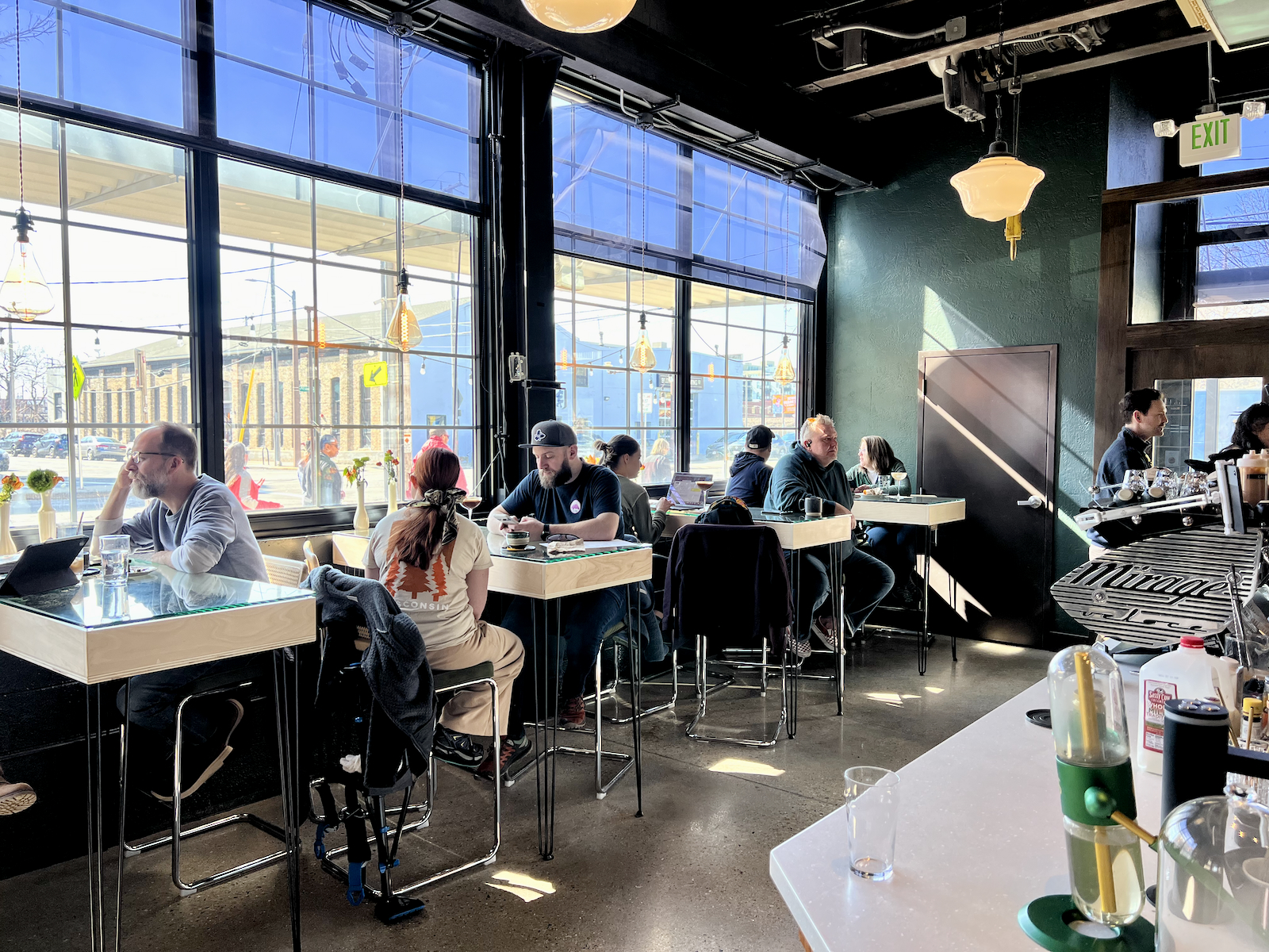 Interior of a contemporary cafe with large windows, multiple customers seated at simple tables, black ceiling with hanging lights, and a green accent wall with an exit sign.