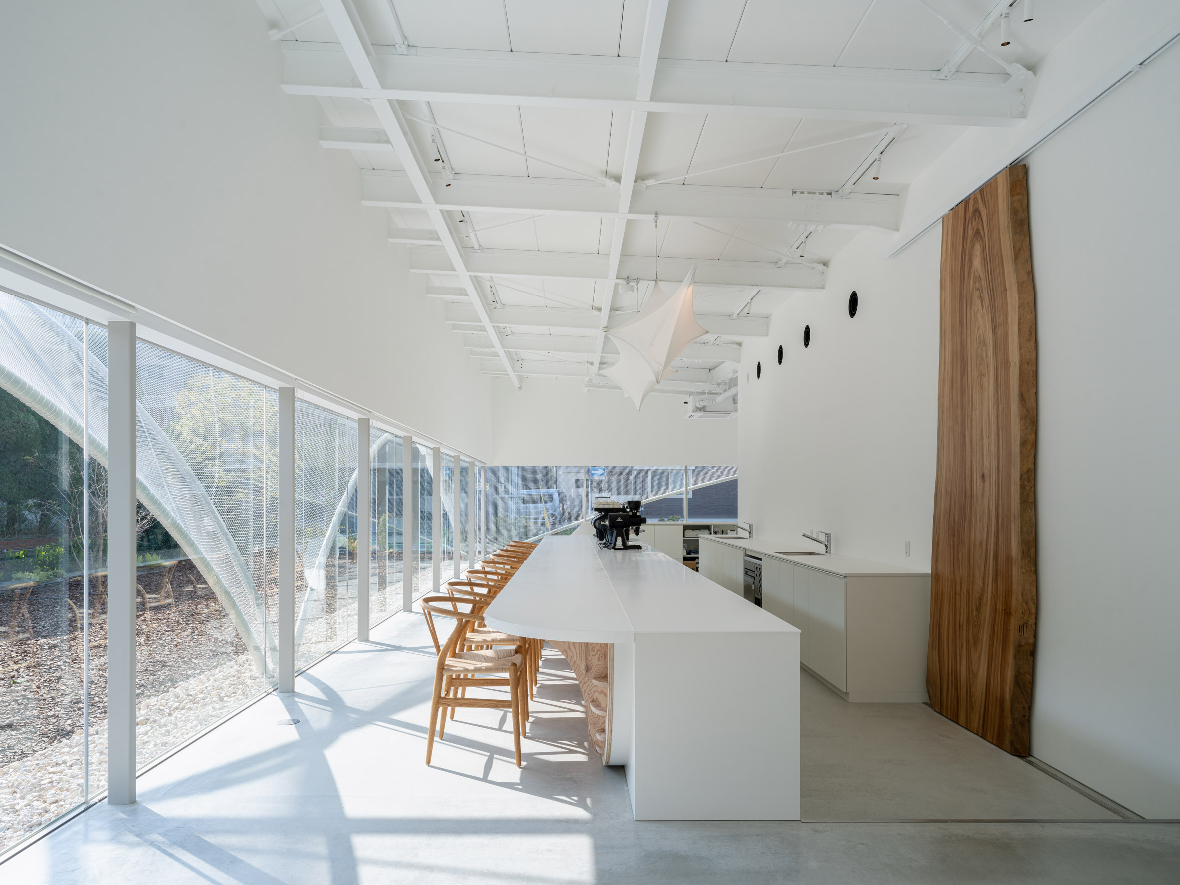 Modern minimalist café interior with white coffered ceiling, long white counter/bar with espresso machine, wooden chairs, and floor-to-ceiling windows