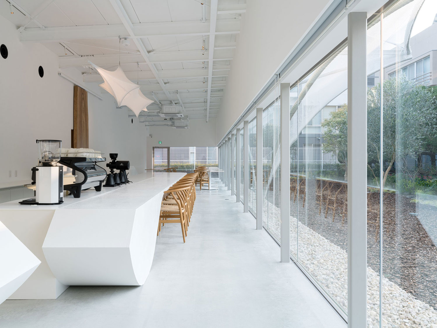 Interior of a modern coffee shop featuring a white curved counter with espresso machine, wooden chairs along a long counter, and extensive glazing on the right side opening to an outdoor garden area.