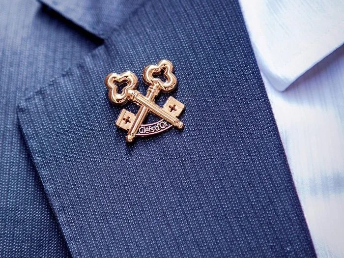 A close-up of the gold crossed-keys Les Clefs d'Or lapel pin, with "Clefs d'Or" visible, worn on a navy suit jacket.