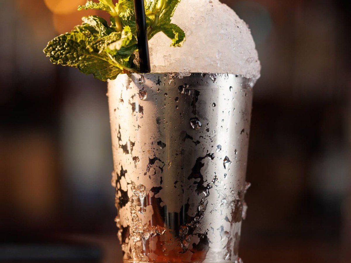 A close-up of a Mint Julep cocktail in a silver metal cup, overflowing with crushed ice and garnished with fresh green mint leaves, with a black