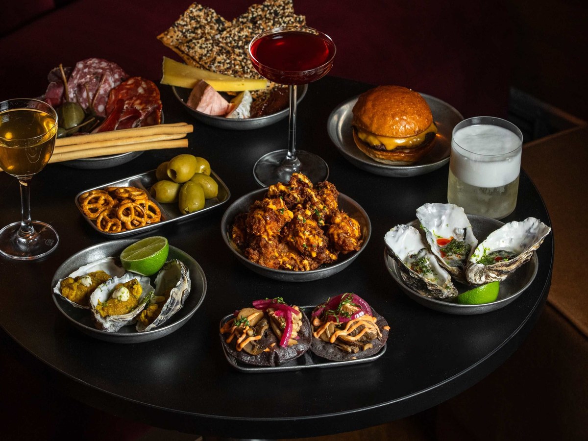 A dark round table is laden with various small plates and drinks, including charcuterie, oysters, fried chicken, a burger, and cocktails.