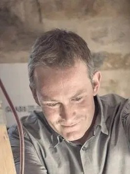 A man with short gray hair wearing a gray shirt concentrates on painting with a brush in his hand.