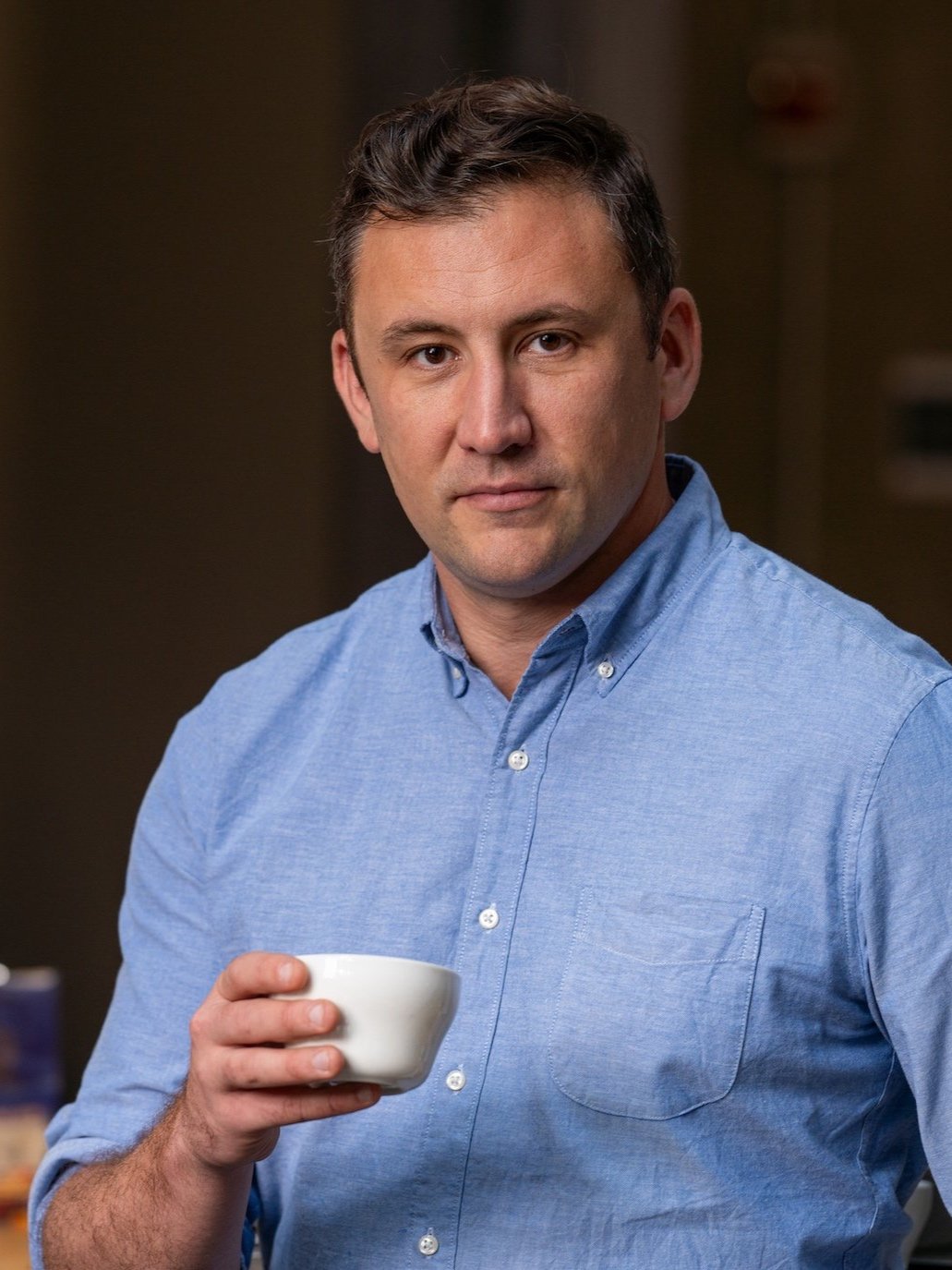 Professional portrait of a man in a light blue button-down shirt holding a white coffee cup, shot in an indoor setting; source page explicitly identif