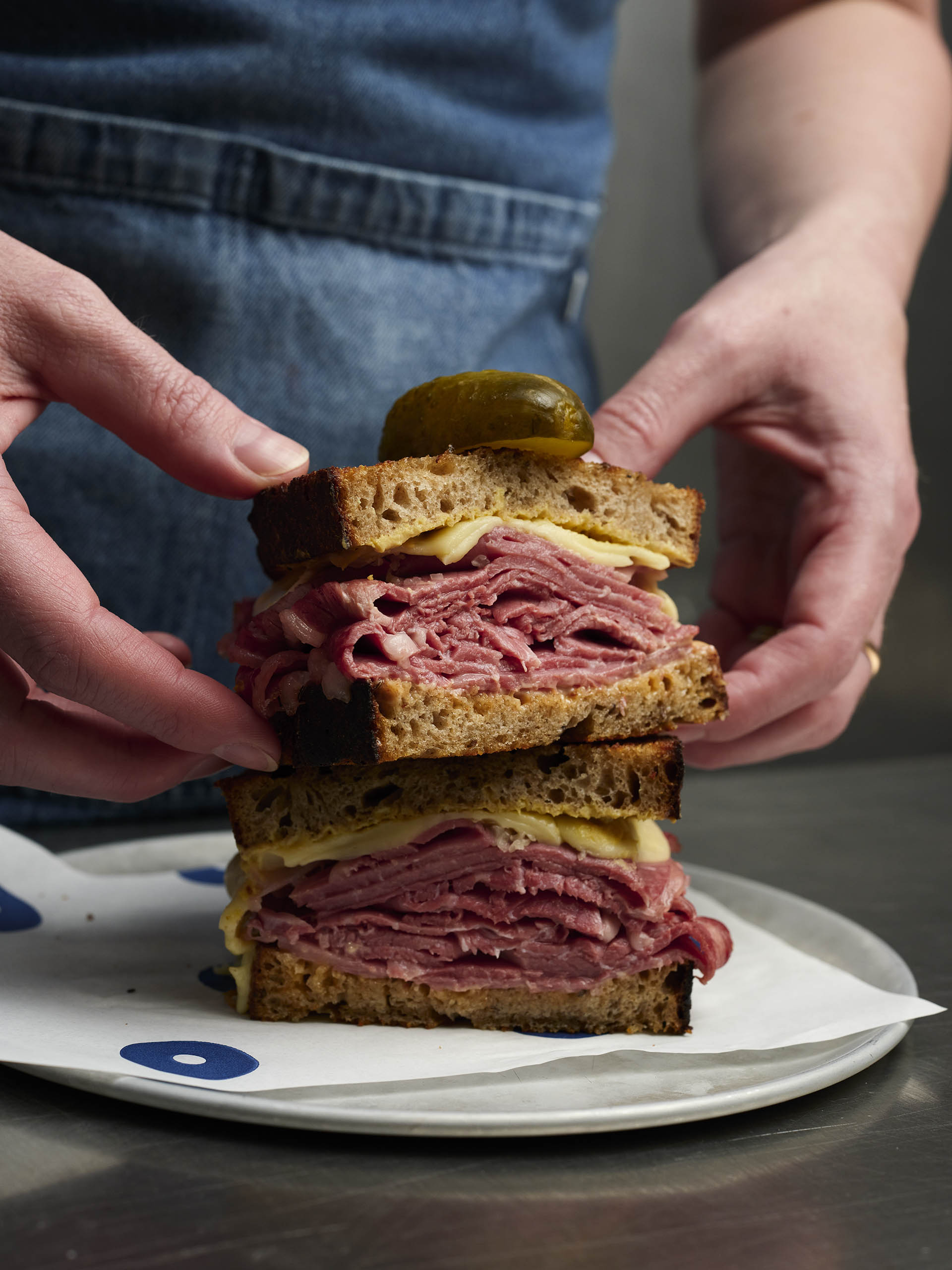 A stacked sandwich with thick slices of pink salt beef, cheese, and a pickle on dark sourdough bread, held by hands in frame