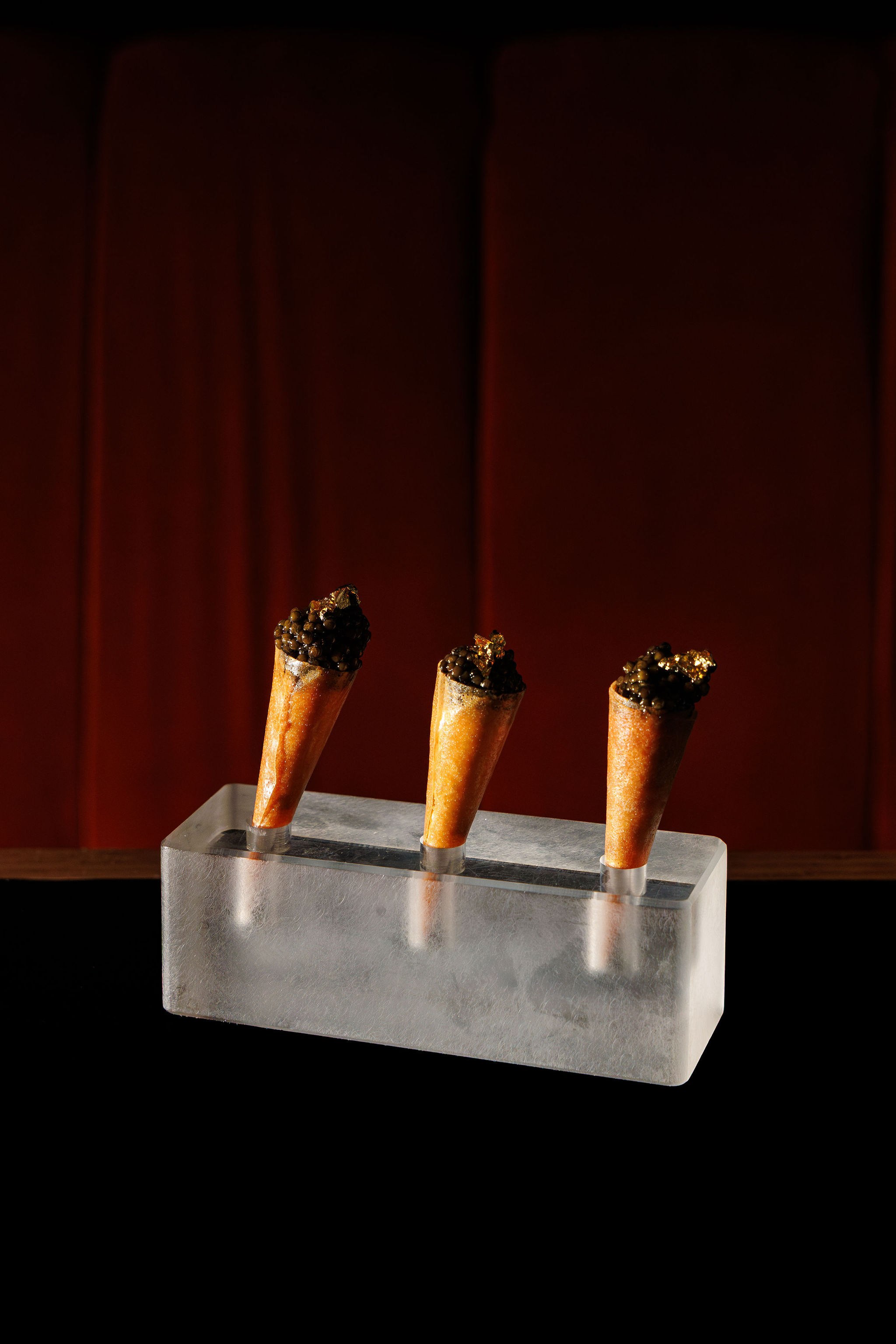 Three burning cigars with wooden bands inserted into holes in a rectangular concrete block, photographed against a dark red curtain background.