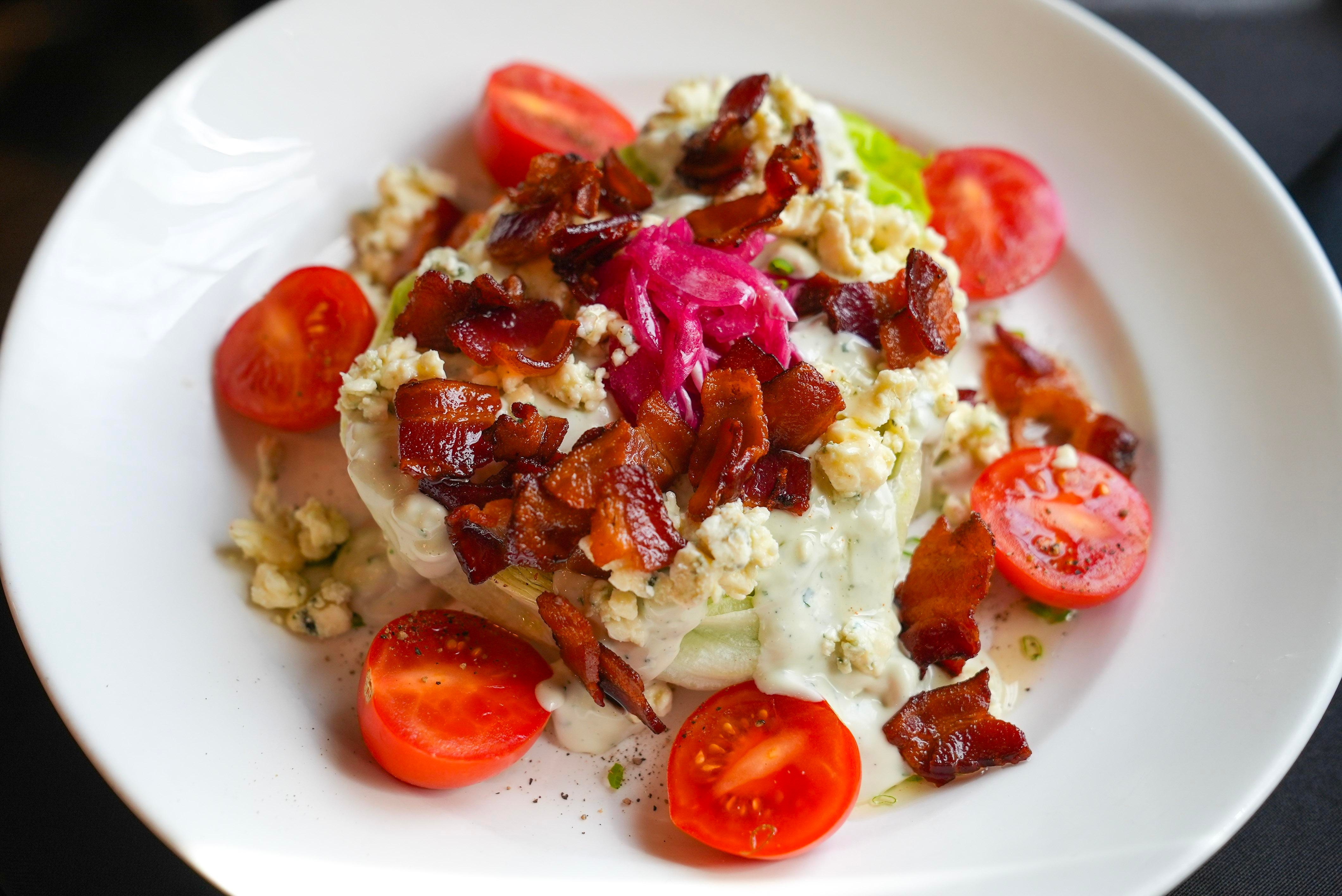 A white plate with burrata cheese topped with bacon, pickled onions, and nuts, surrounded by sliced tomatoes.