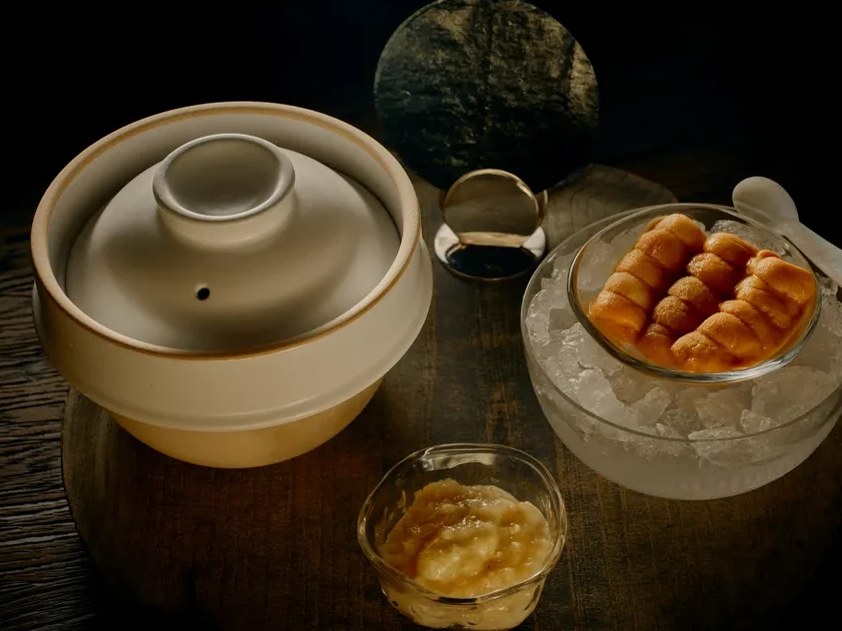 Inside a dark lair for drinking, a ceramic pot and cocktail presentation on ice sit on a wooden table.