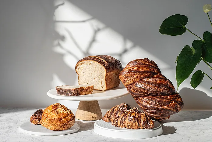 Artisan bakery display showing fresh sourdough loaves, braided bread, croissants, and pastries arranged on white cake stands and marble surface; the s