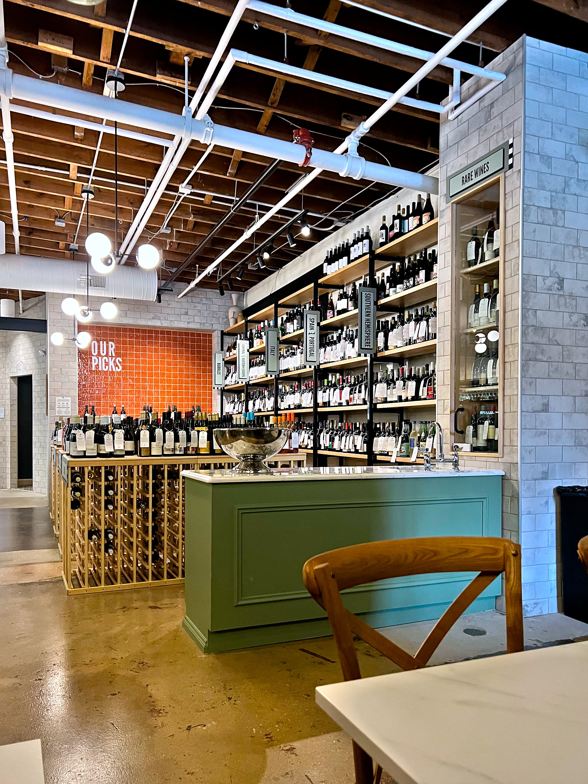 Wine shop counter with green facade and wooden wine storage below; shelves of bottles and 'OUR PICKS' signage visible behind