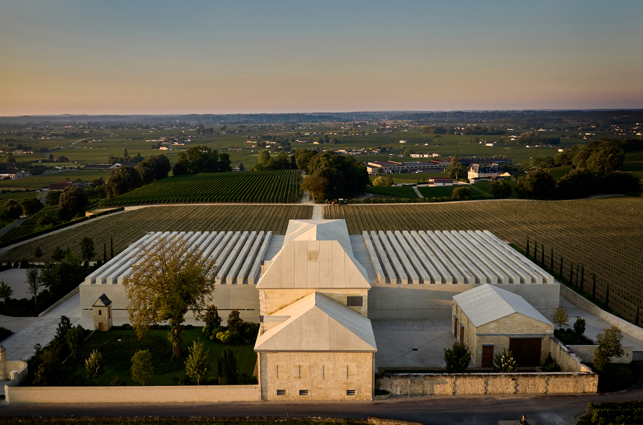 Modern winery with white-roofed barrel halls overlooking striped vineyard rows in rolling green landscape