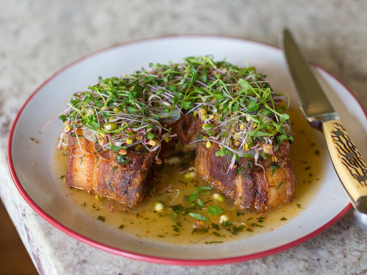Two pieces of crispy pork belly, topped with a vibrant mix of green and purple microgreens, sit in a shallow pool of sauce on a white plate with a