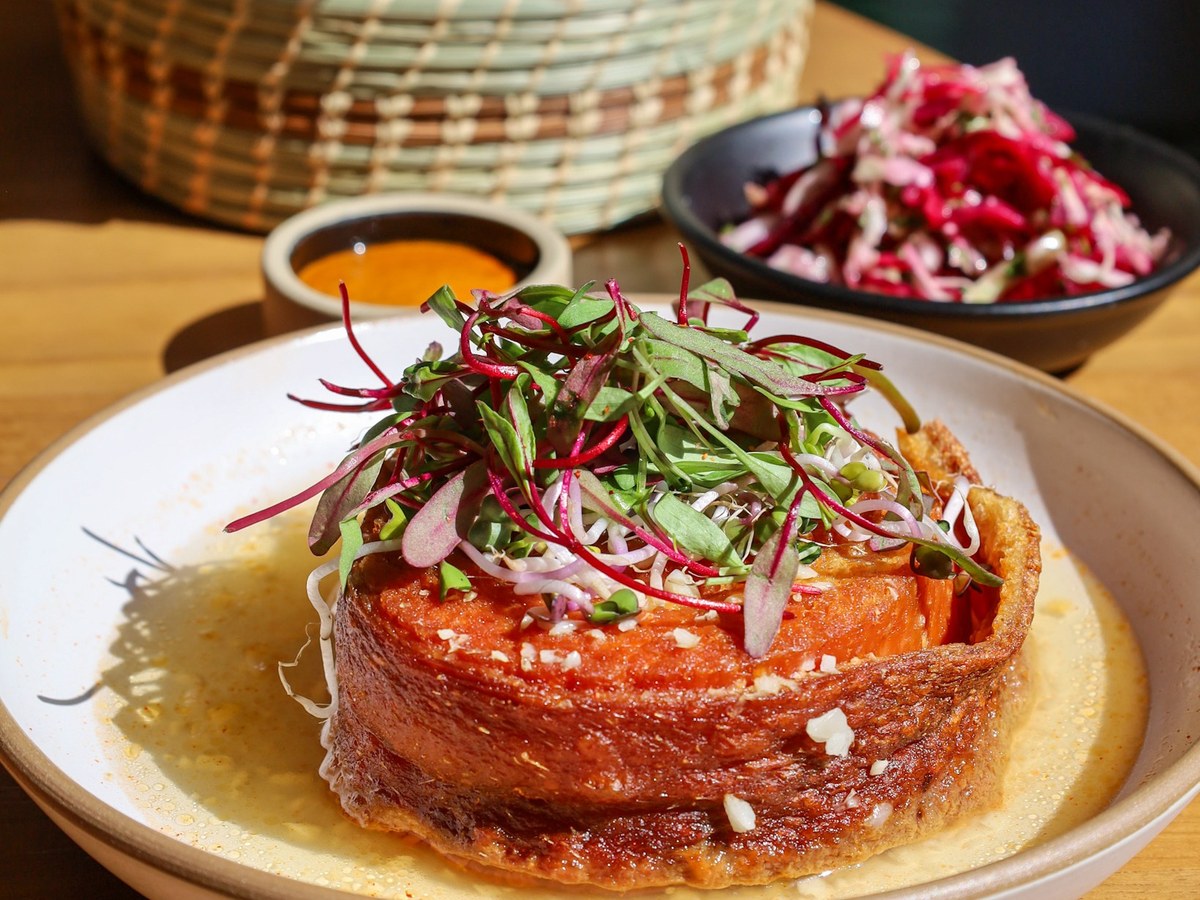 A close-up of a dish from Broken Spanish Comedor, featuring a cylindrical, crispy-edged item in a shallow pool of sauce, topped with fresh