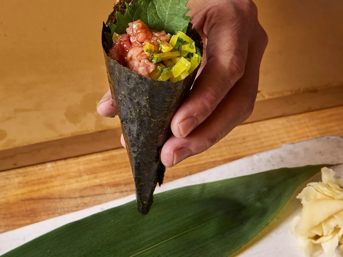 A hand holds a temaki (hand roll) sushi cone wrapped in nori, filled with minced tuna, scallions, and shiso leaf, presented over a bamboo-leaf-lined p