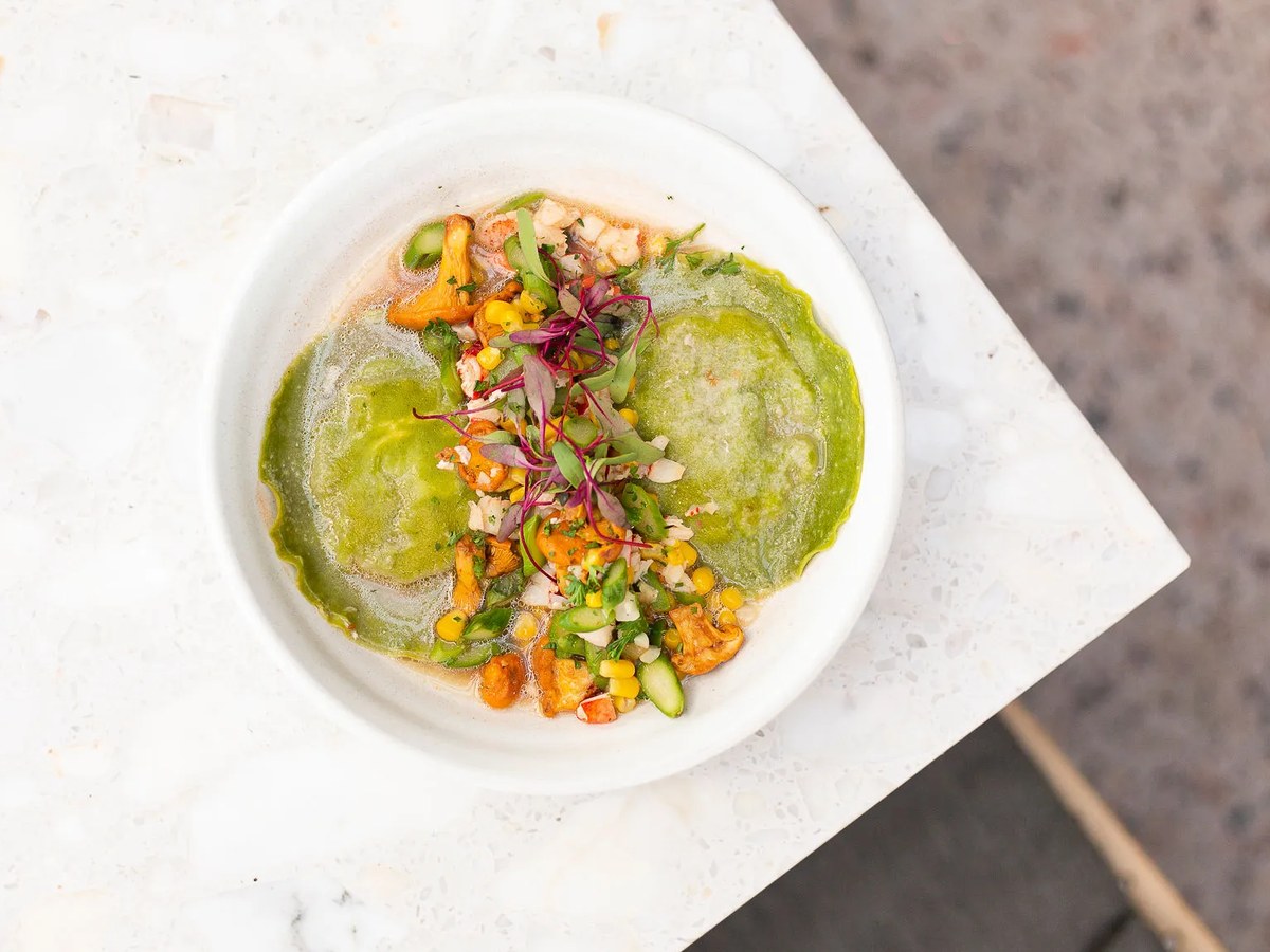 An overhead editorial shot of a plated fine-dining dish featuring two green ravioli in a light broth, garnished with chanterelle mushrooms, corn, micr