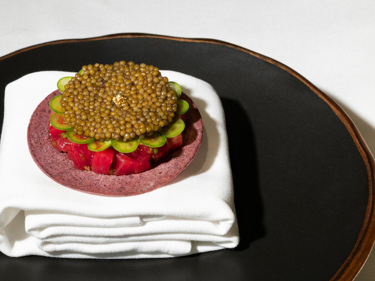 An elegantly plated fine-dining dish featuring caviar atop thinly sliced vegetables arranged in a flower pattern on a purple base, presented on a folded white napkin