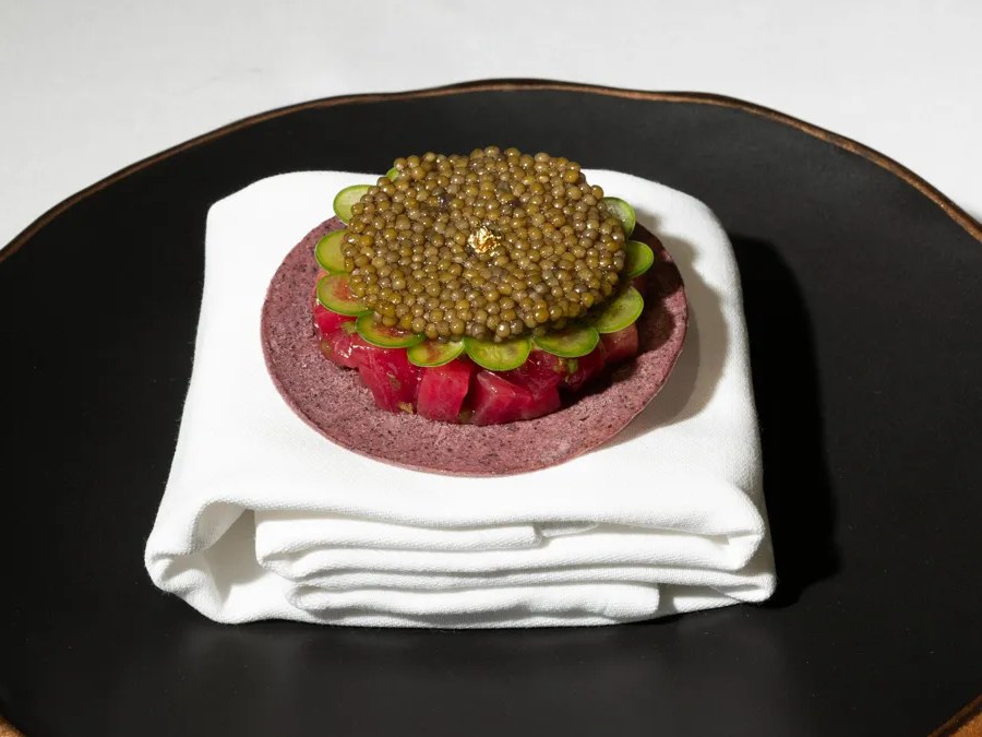 An elegantly plated tasting menu course featuring caviar atop sliced green vegetables and diced red fish, presented on a purple disc on a white napkin