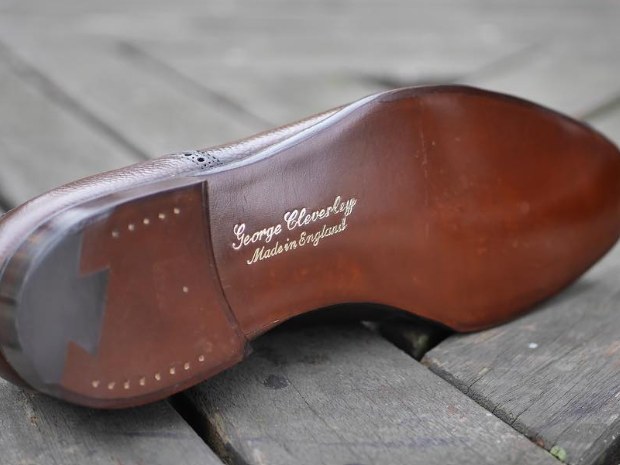The brown leather sole of a George Cleverley shoe, with 'George Cleverley / Made in England' stamped in gold, rests on wooden planks.