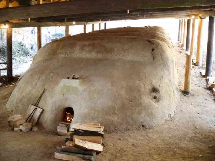 A large, light brown, mud-built climbing kiln with a small arched opening at its base, where a fire glows, is sheltered under a wooden roof structure.
