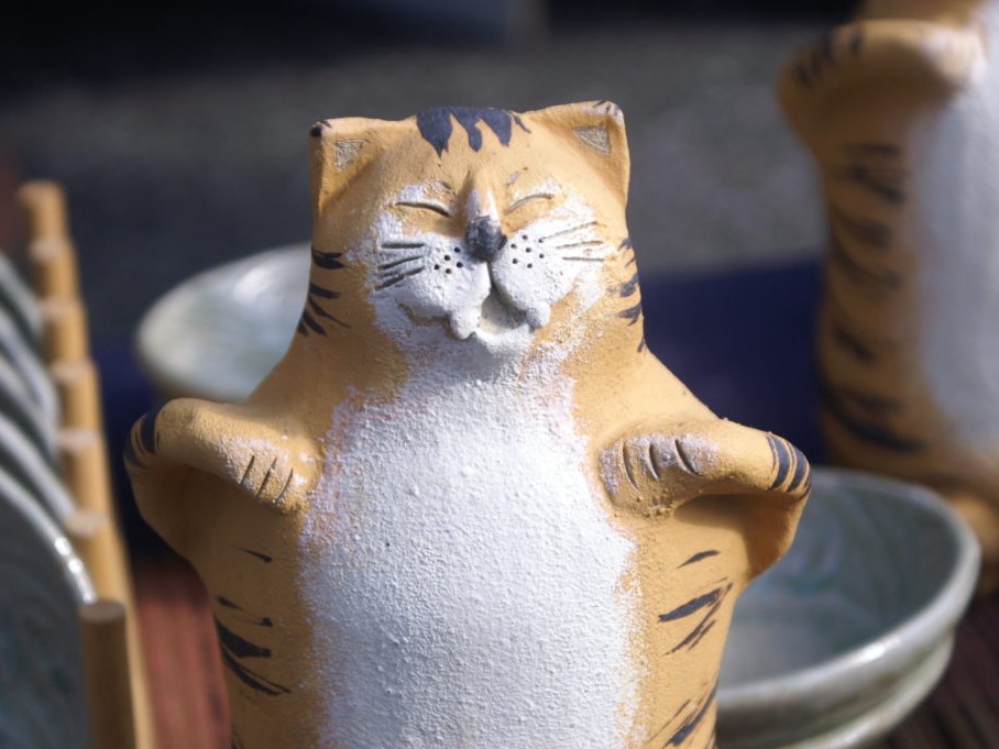 A ceramic cat figurine, orange and white with black stripes, smiles with closed eyes, surrounded by pottery bowls and dishes.