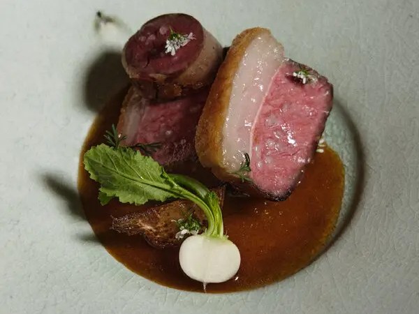 A white plate holds sliced pink meat, a small white turnip with green leaves, and dark brown sauce.