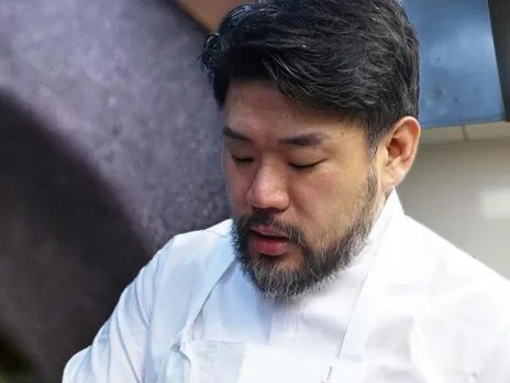 A chef with dark hair and a beard wearing a white chef's coat looks down while working.