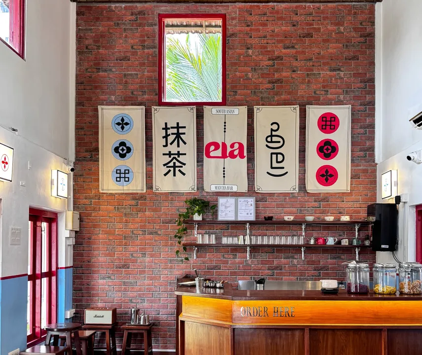 Interior of a matcha cafe showing a brick wall with decorative banners, a service counter with 'ORDER HERE' signage, shelving with cups and equipment,