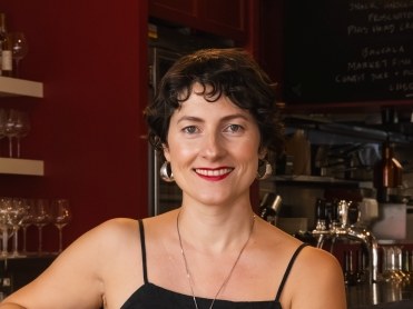 A smiling woman with curly dark hair wearing a black sleeveless top and necklace, photographed indoors at what looks like a wine bar or restaurant.