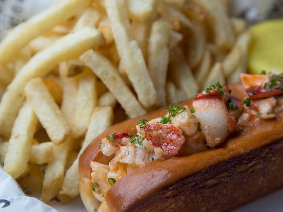 A close-up of a lobster roll with chunks of lobster meat and chives on a toasted bun, served with a side of golden french fries and a yellow lemon