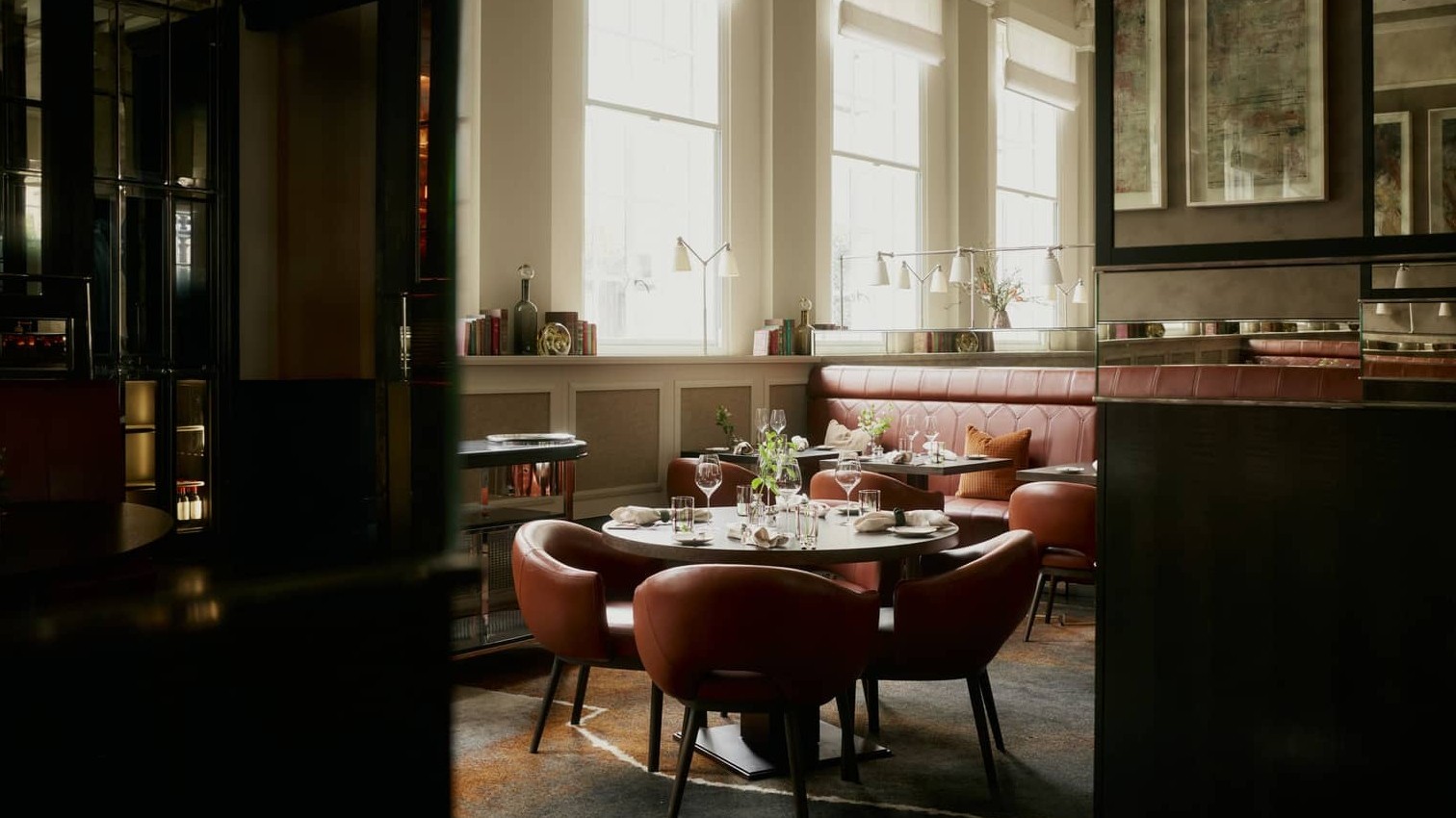 An elegant dining room with brown leather chairs, banquettes, and round tables set with glasses, next to tall windows.