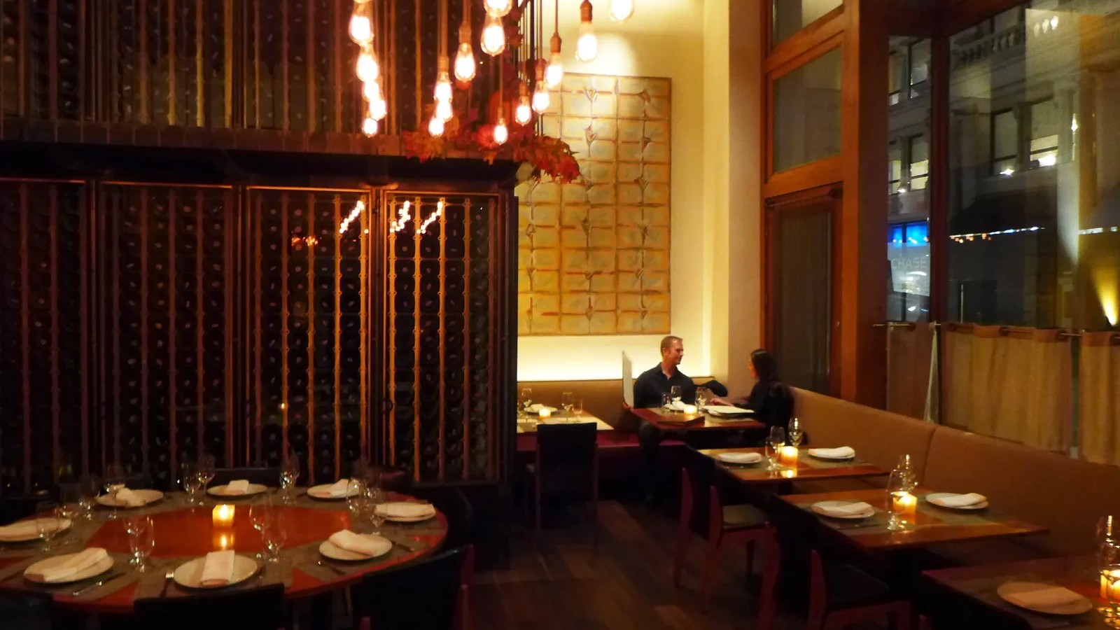 A warmly lit fine-dining restaurant interior with dark wood wine storage walls, Edison-bulb pendant lighting, and tables set for service.