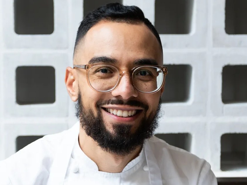 A smiling man with a beard and clear-framed glasses wearing a white chef's coat and apron, seated in front of a decorative white breeze-block wall, id