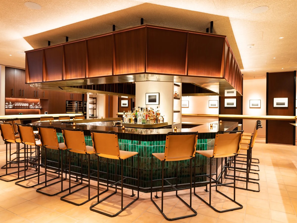 A polished bar interior featuring a curved counter clad in deep green tile with tan leather barstools, warm wood overhead canopy, and a full liquor di