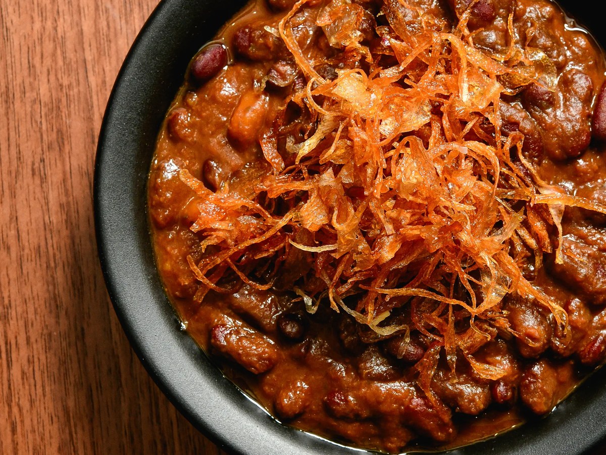 A black bowl containing a rich bean and meat stew topped with crispy fried shallots or onions, served on a wooden surface, consistent with a halal La