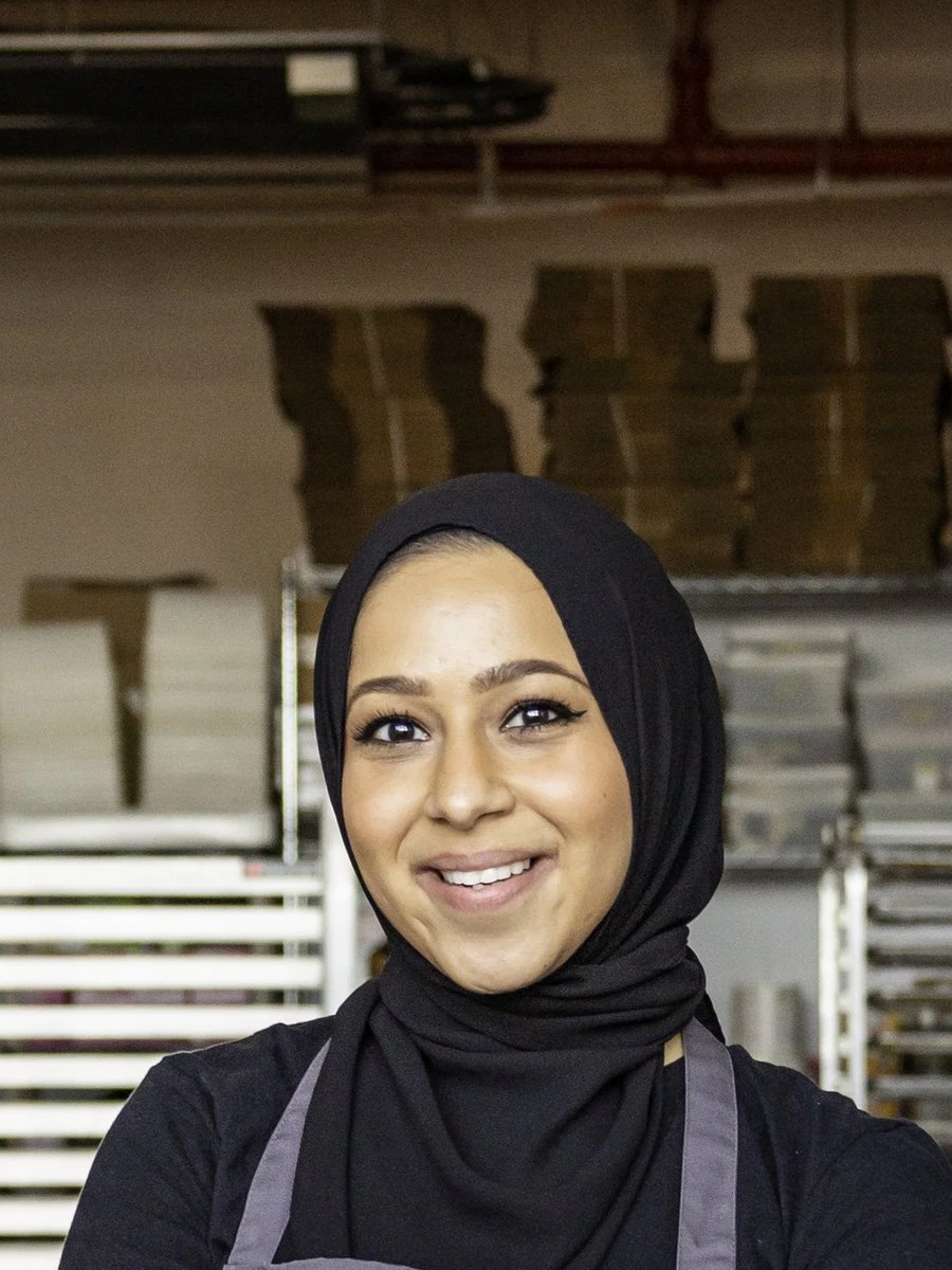 A woman wearing a black hijab and grey apron stands with arms crossed in what appears to be a commercial bakery kitchen; identified as Sumaiya Bangee 