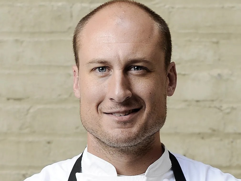 A portrait of chef Dave Beran, a bald man with blue eyes and a slight smile, wearing a white chef's jacket and a black apron against a light-colored