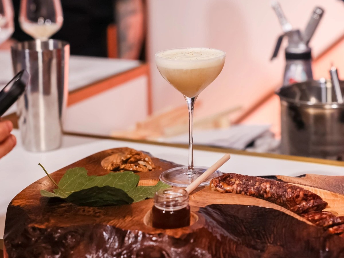A frothy cream-colored cocktail in a coupe glass sits atop a rustic wooden charcuterie board with cured meats, walnuts, fig leaves, and a small jar of
