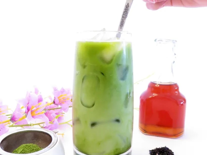 A tall glass containing a layered green matcha drink with visible tea leaves, being stirred by hand; the source page explicitly attests this is 'earl 