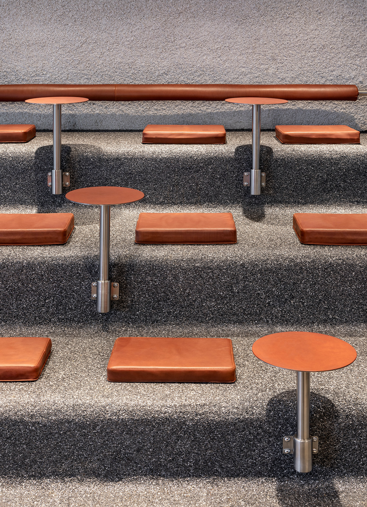 Tiered concrete seating area with orange/terracotta leather cushions and small round tables on metal poles, showing the multi-level communal seating d