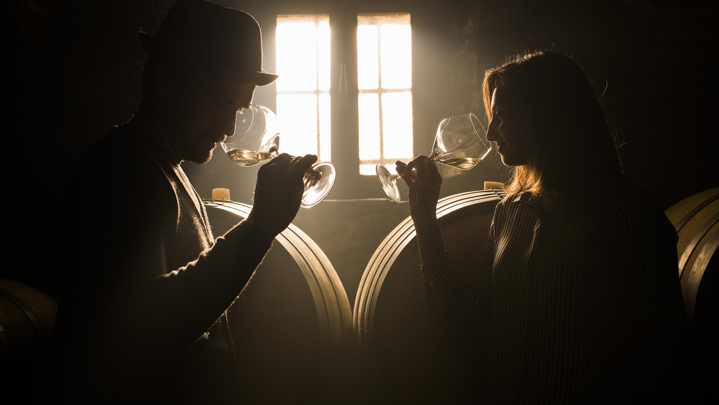 Two people holding clear wine glasses against bright window light, pouring white wine in warm-toned interior