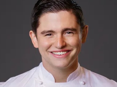 A young man with dark hair, wearing a white chef's coat with green embroidered details, smiles at the camera against a gray background.