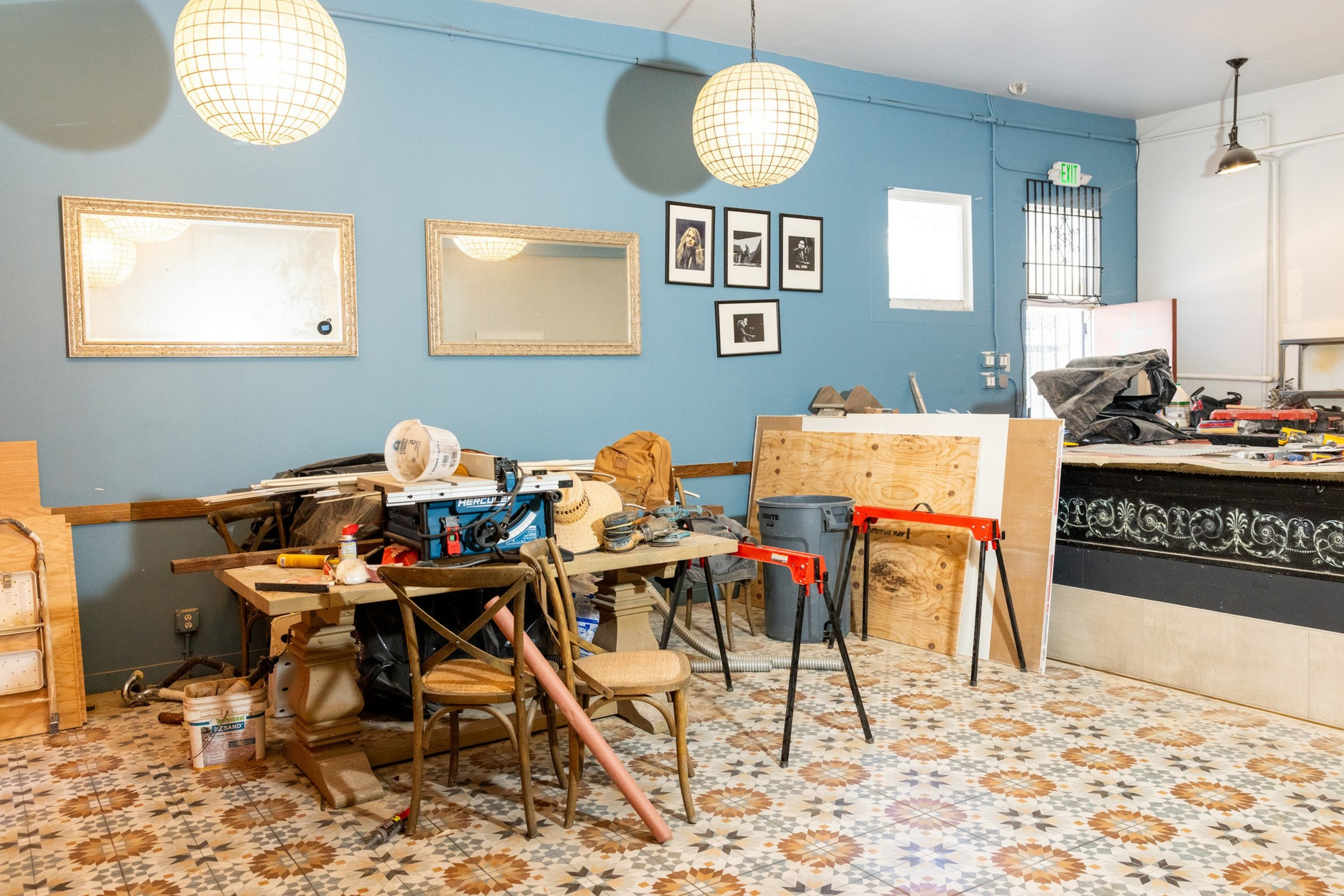 A room undergoing renovation with construction materials, tools, work tables, patterned tile flooring, two large ornate mirrors, and globe pendant lig