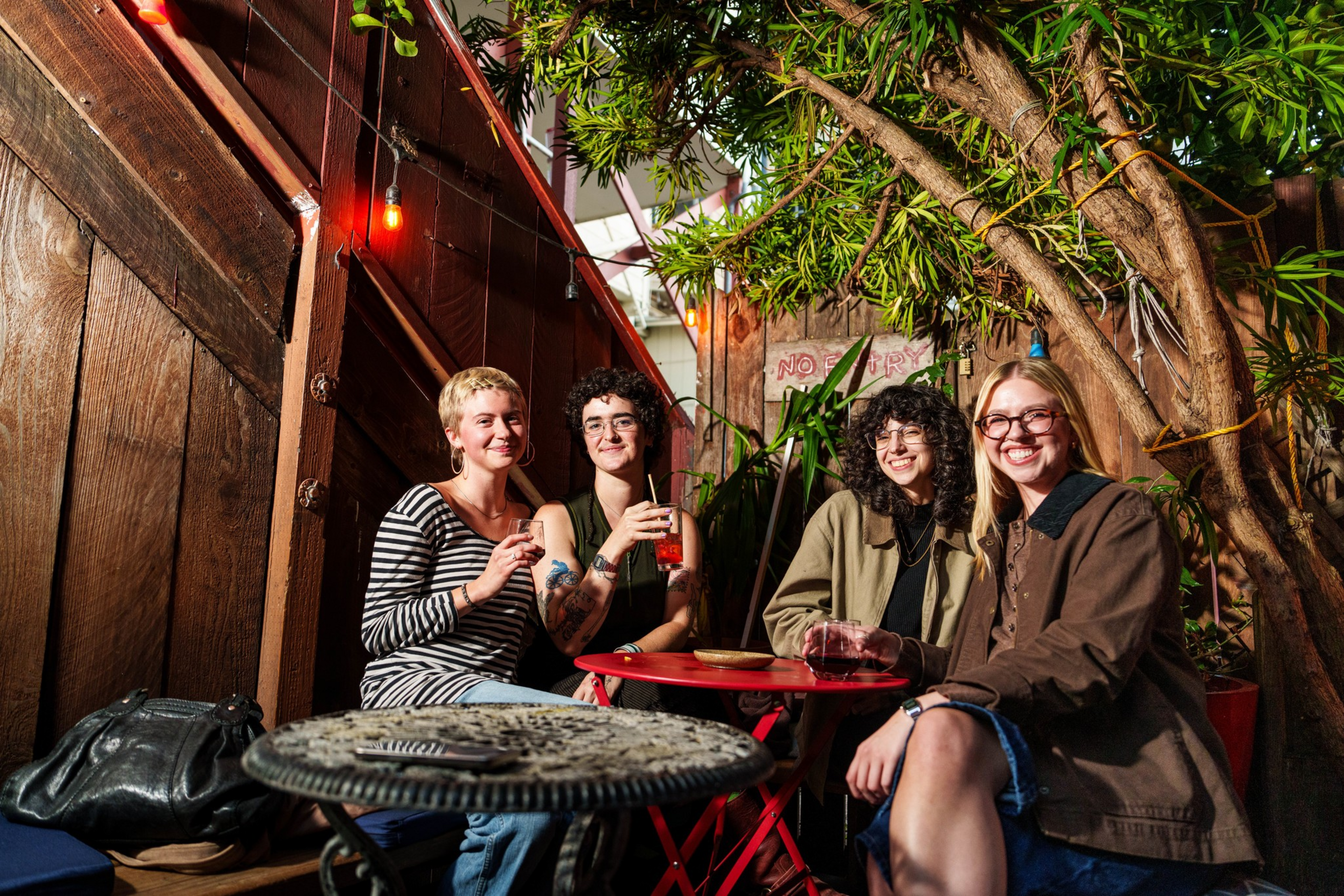 Four friends sit around a small red table in an outdoor patio setting with warm string lights and greenery