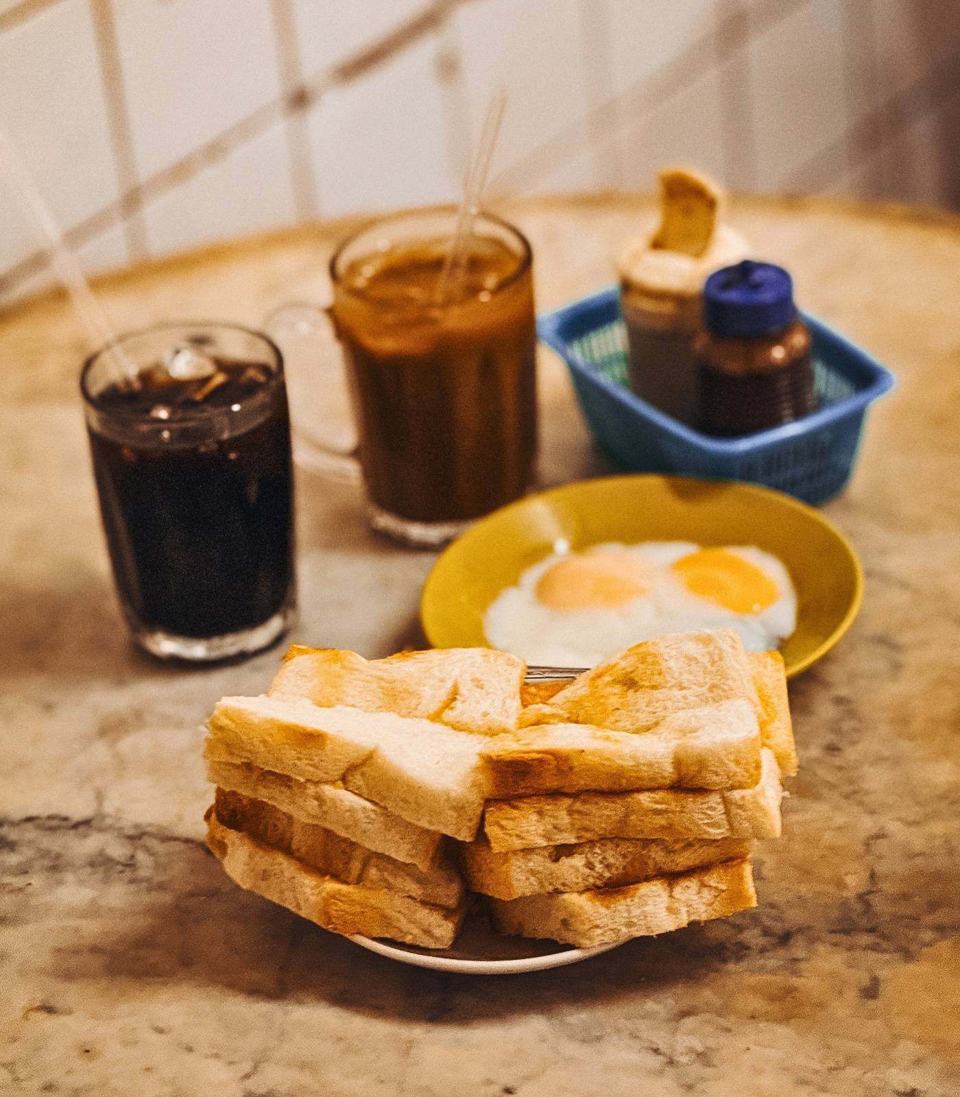 Traditional Nanyang breakfast setup showing kaya toast slices stacked on a plate, two fried eggs on a yellow plate, and dark beverages including what
