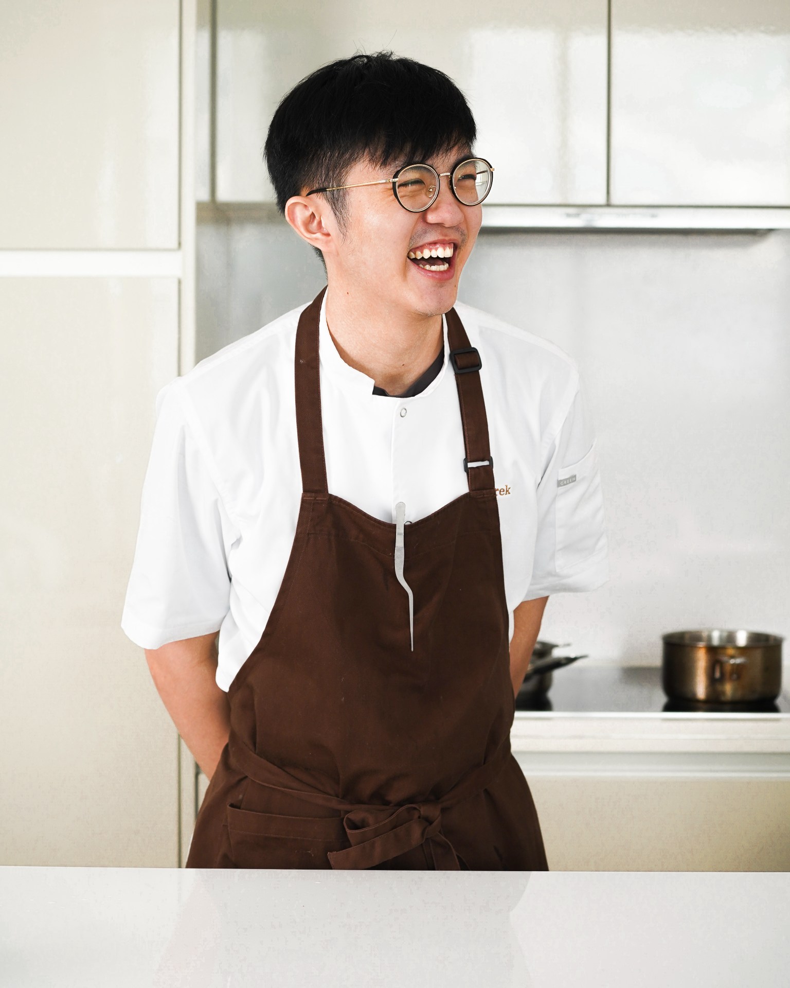 A young man with short black hair wearing glasses, a white t-shirt, and brown apron stands in a bright kitchen with a warm, genuine smile.