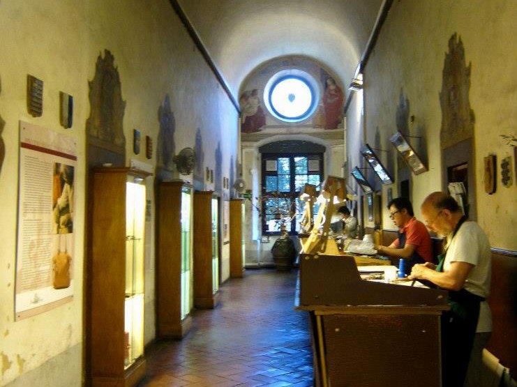 A long, vaulted corridor with terracotta floors and frescoed walls, where two men work at a wooden bench in a leather workshop.