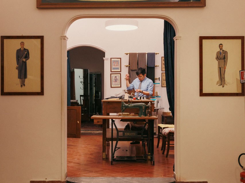 A tailor works at a Borletti sewing machine inside the Liverano & Liverano atelier, with a prominent 'LIVERANO' sign above the arched entrance.