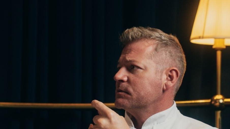 A man with blonde hair and facial hair wearing a white shirt, photographed in profile against a dark interior setting with a lamp visible in the background.
