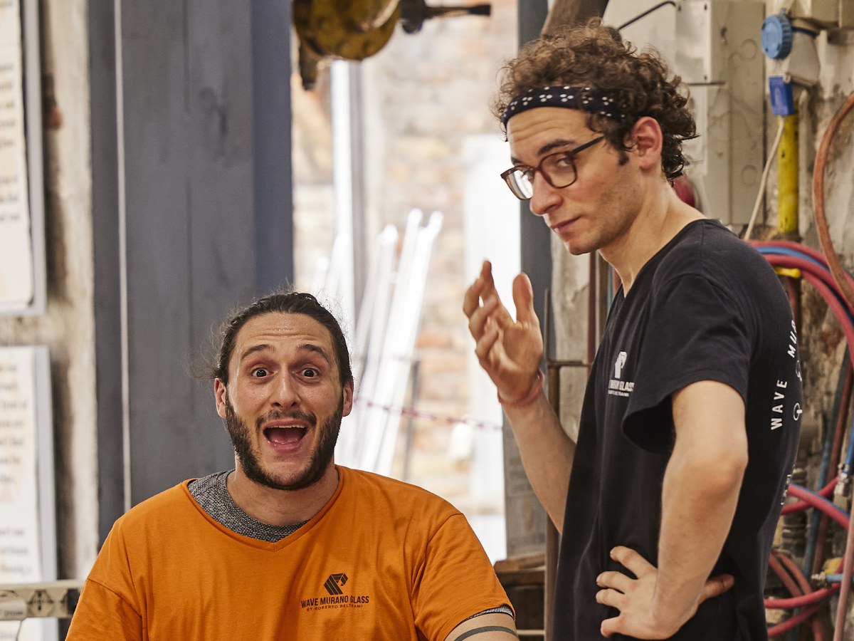 A glassblower in an orange shirt works molten glass on a bench, while a colleague and two visitors observe in a workshop.